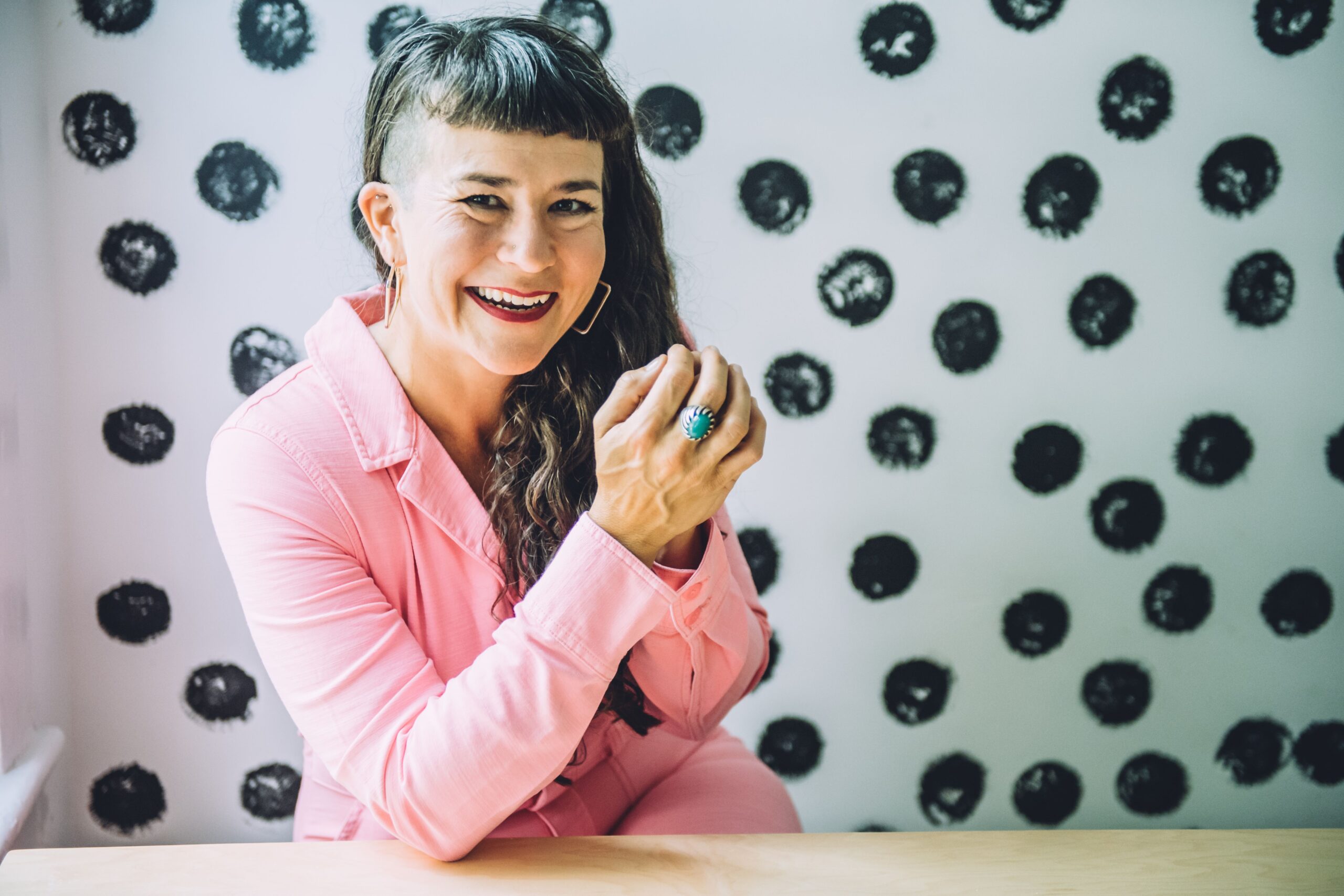 Photo of life coach Beatriz Victoria Albina in a pink jumpsuit smiling in front of a white wall painted with dark polka dots.